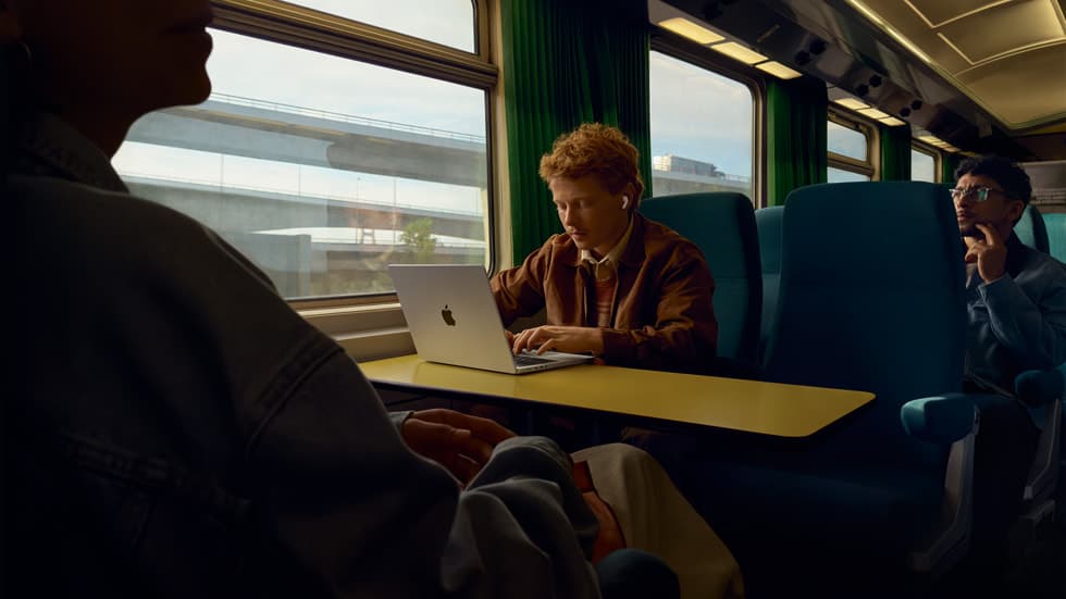 Person med AirPods Pro 3 bruker en MacBook paa et tog, mens medreisende sitter i bakgrunnen