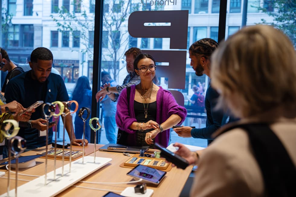 Kunde prøver Apple Watch ved et bord med Apple Watch-modeller i en Apple Store, mens en ansatt hjelper til