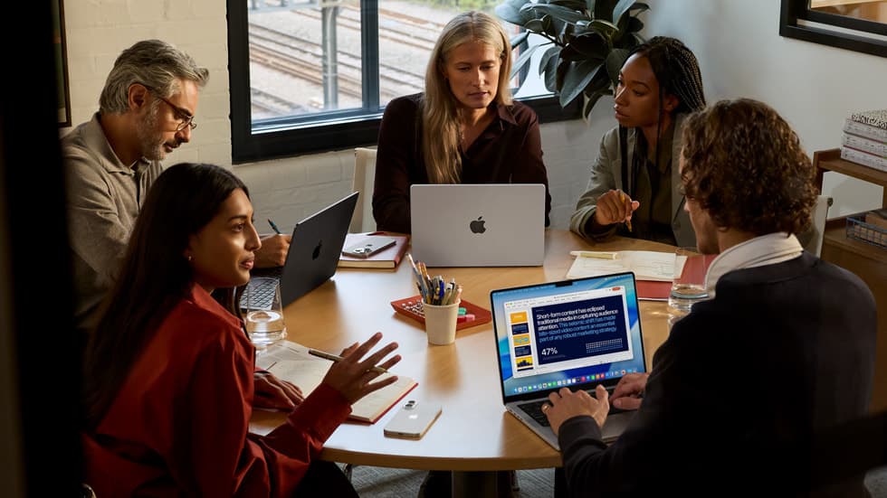 Flere personer i et arbeidsmøte rundt et bord, der flere bruker MacBook Pro og en presentasjon med dashbord vises på skjermen i forgrunnen