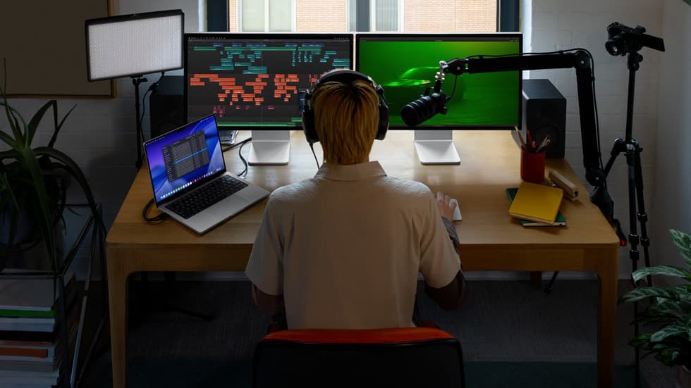 En videoskaper redigerer ved et skrivebord med MacBook Pro og to Apple-skjermer som kjorer Final Cut Pro og Logic Pro