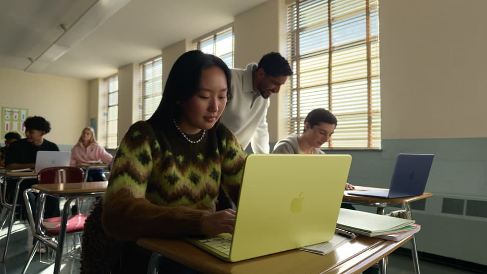 Elev bruker en sitrongul MacBook Neo i et klasserom mens en lærer hjelper, med en medelev og en indigoblå MacBook Neo i bakgrunnen