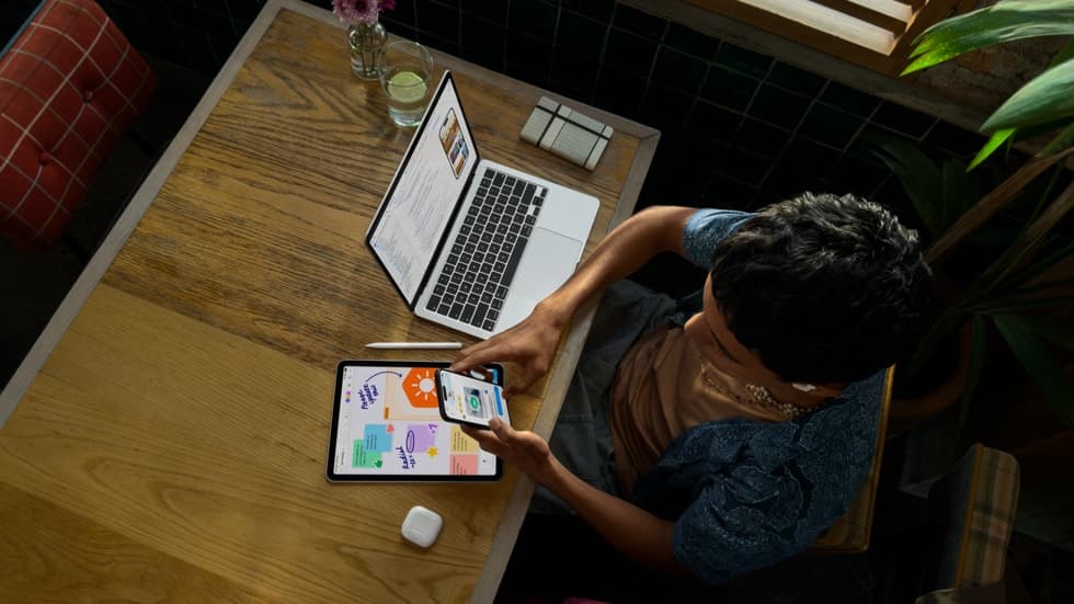 En person vises ovenfra. Vedkommende holder en iPhone og jobber på en MacBook Pro ved et bord. Det vises kode på skjermen på MacBook Pro. Ved siden av ligger iPad, Apple Pencil og AirPods.
